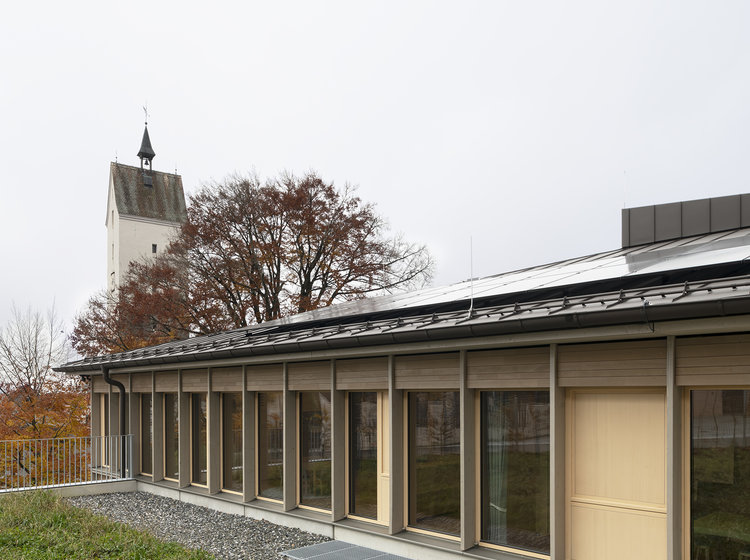Modernes Flachdachgebäude mit großer Fensterfront und Holzfassade, umgeben von Grünfläche; im Hintergrund ein Kirchturm und herbstliche Bäume.