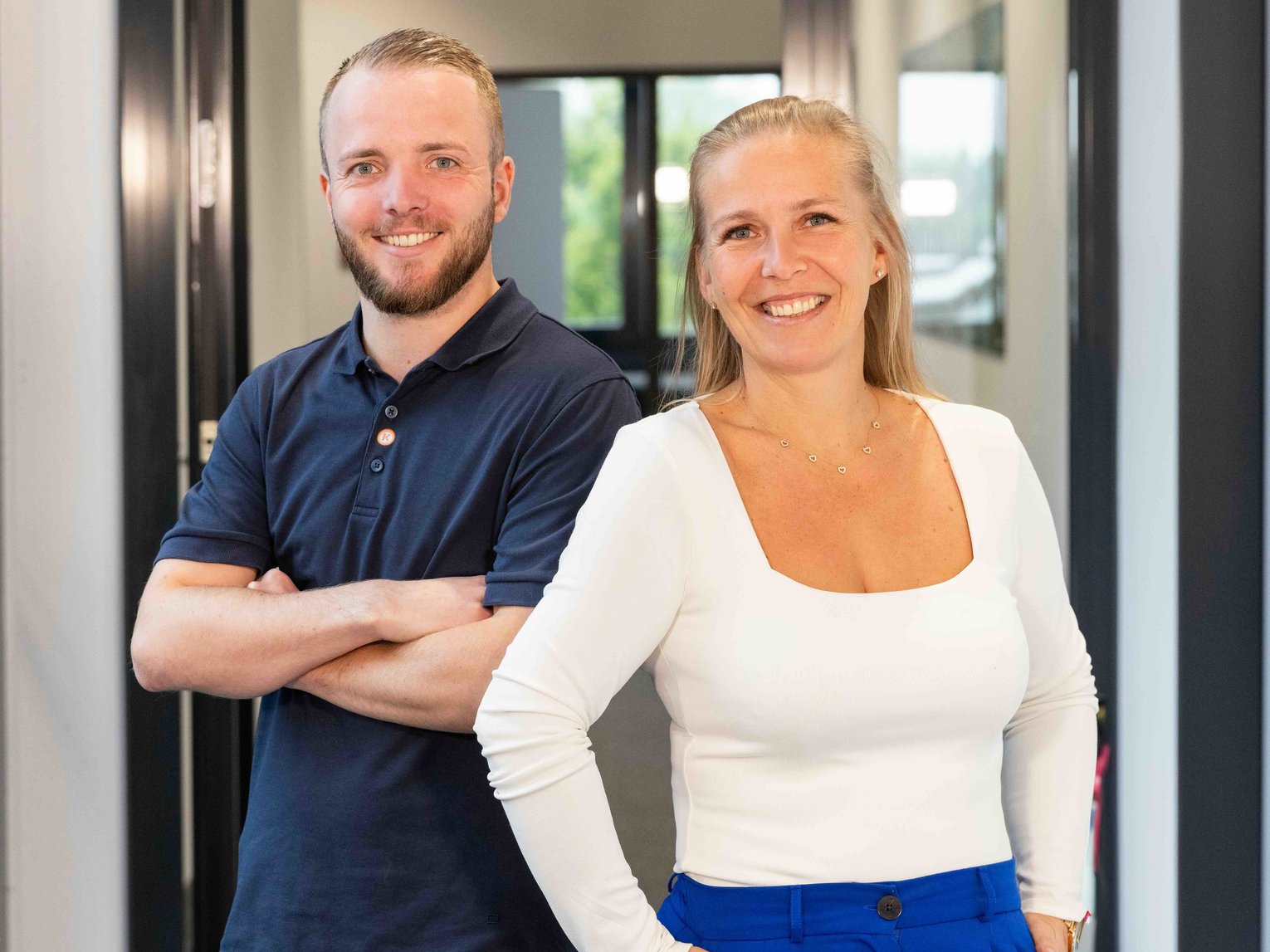 Zwei lächelnde Mitarbeitende stehen in einem modernen Büroflur nebeneinander, die Arme bzw. Hände selbstbewusst positioniert, und blicken freundlich in die Kamera.