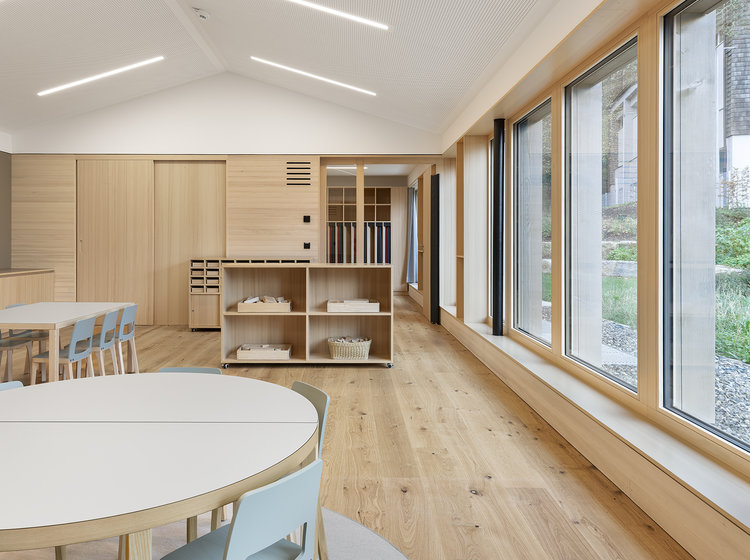 Heller Gemeinschaftsraum im Kindergarten St. Vincenz mit runden Tischen, Holzfußboden und großer Fensterfront mit Blick ins Grüne.