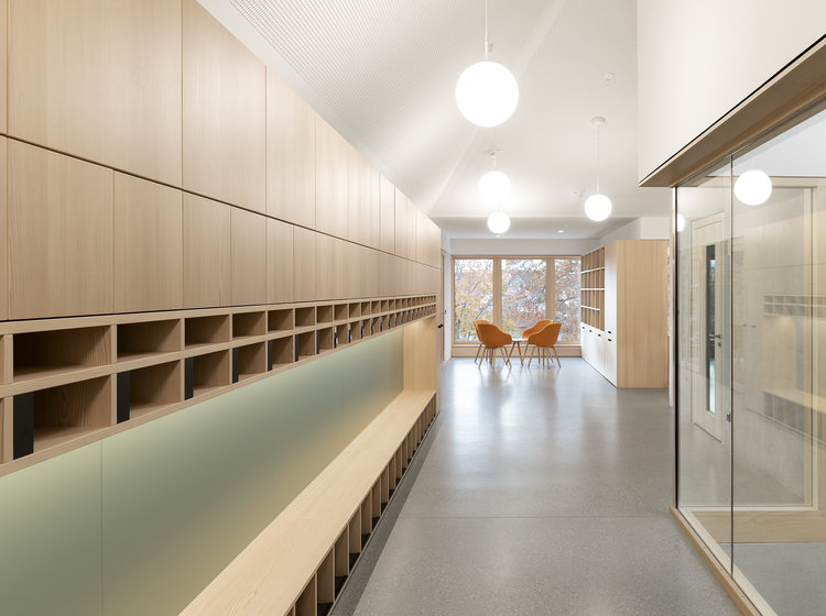 Heller Flur mit Holzregalen und Sitzbank, modernen Glastrennwänden und runden Deckenleuchten; im Hintergrund zwei orangefarbene Stühle.