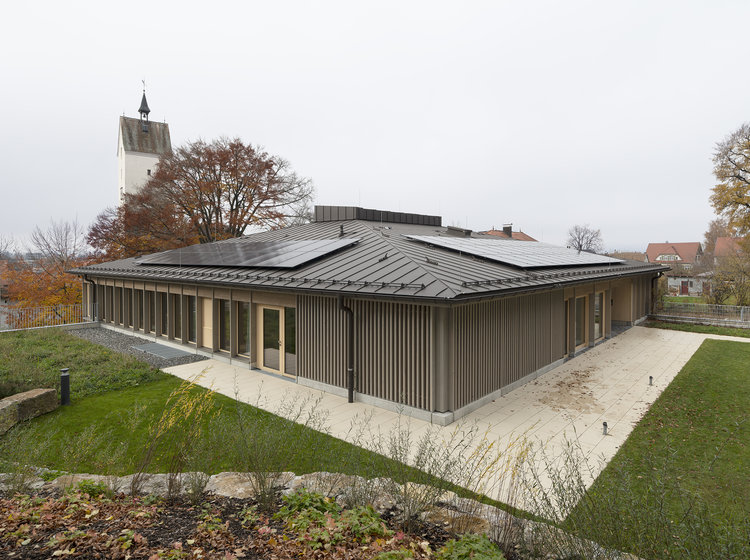 Energieeffizienter Neubau mit dunkler Holzfassade, Flachdach und Solarpanels; umgeben von Rasenflächen, im Hintergrund ein Kirchturm und Bäume.