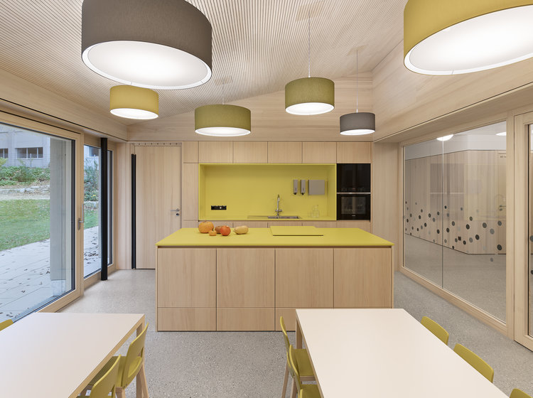 Moderne Küche mit gelber Rückwand, Kochinsel und großen Hängelampen; angrenzender Essbereich mit weißen Tischen und grün-gelben Stühlen.